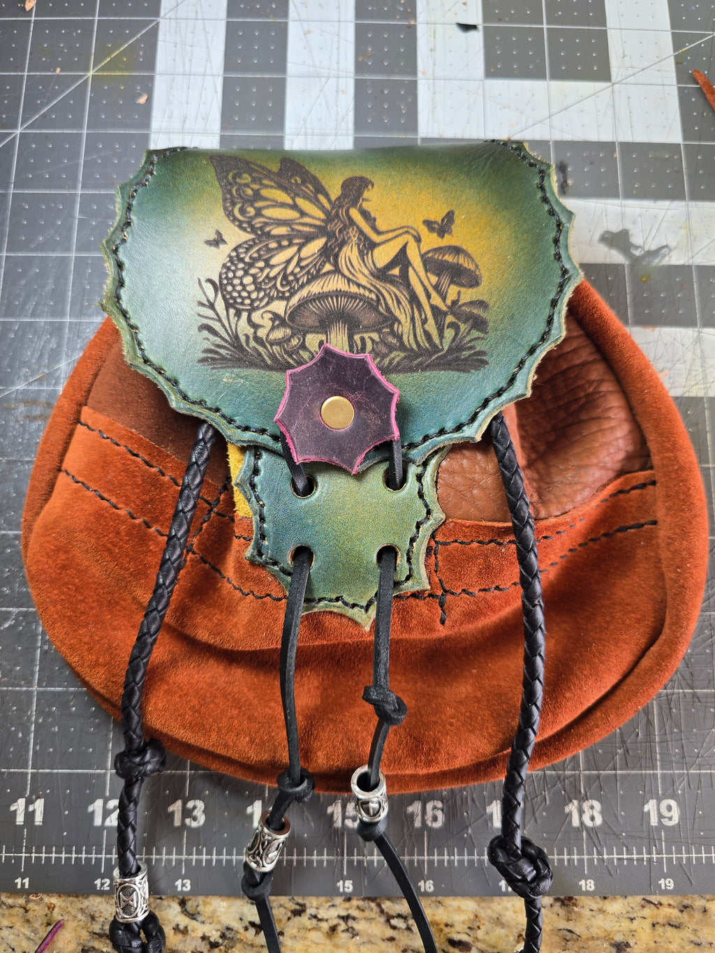 Fae-Stitch Satchel detail showing laser-engraved fairy flap, patchwork buffalo leather body, and black heavy-duty stitching — shown on cutting mat for scale