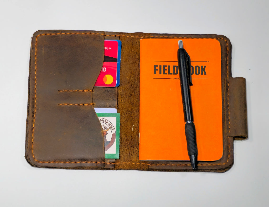 Brown leather mini journal with orange field notebook, pen, and cards on a white background